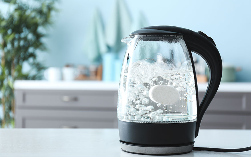 Une bouilloire en verre avec une base noire est sur une table blanche. L'eau à l'intérieur bout, avec des bulles visibles.