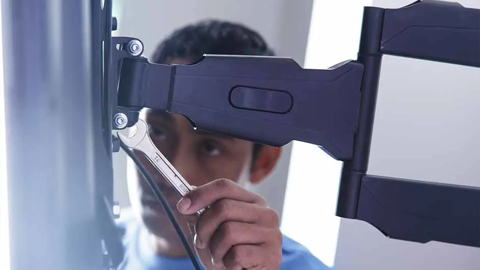 Un support TV noir est fixé au mur avec une clé à molette argentée ajustant les boulons, sur un fond blanc neutre.