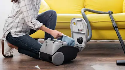 Een stofzuiger op een houten vloer, met een gele sofa op de achtergrond. Iemand verwijdert een blauwe zak.