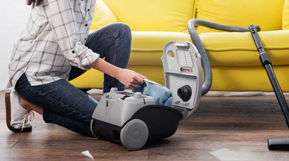 Une personne remplace le sac dans un aspirateur gris et noir sur un plancher en bois, près d'un divan jaune.