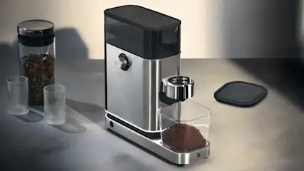 Un moulin à café moderne en acier est posé sur un comptoir, avec du café moulu. À côté se trouvent un pot de grains, deux doseurs et un couvercle.