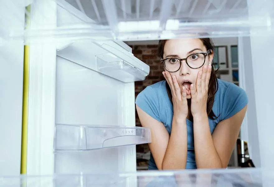 Een open, lege, witte koelkast met transparante schappen is te zien, met op de achtergrond een bakstenen muur en kasten.