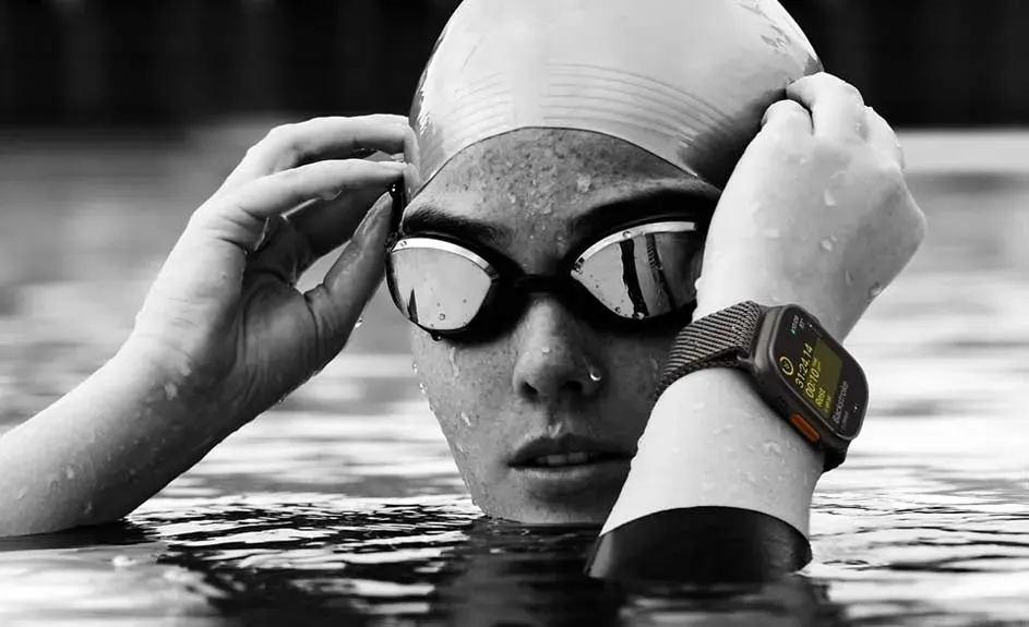 Une personne avec un bonnet de bain et les bras mouillés dans l’eau ajuste ses lunettes de natation, portant une montre connectée affichant des infos sportives.