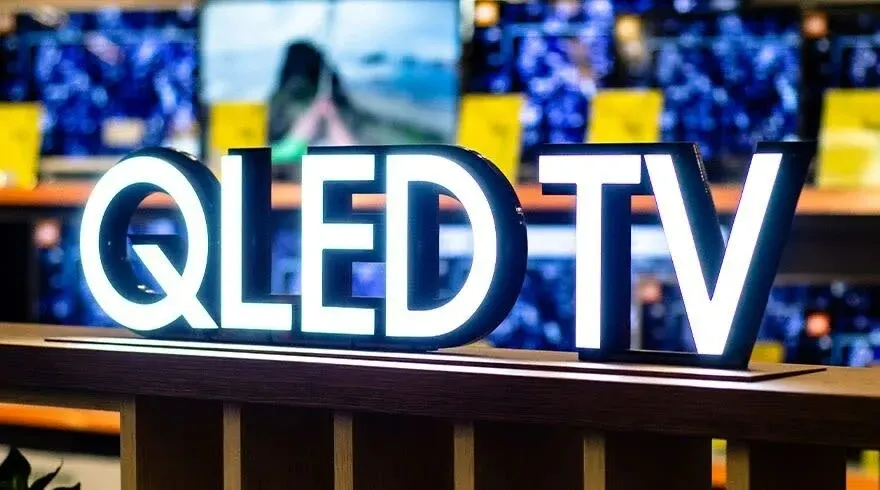 Gros plan sur un signe "QLED TV" lumineux sur une étagère en bois, avec des écrans flous en arrière-plan.