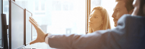 Twee computerschermen staan voor een raam. Een hand wijst naar het scherm, met mensen op de achtergrond.