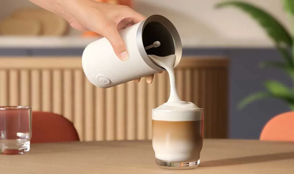 Une main verse du lait chaud depuis un mousseur blanc dans un verre de latte, formant une épaisse couche de mousse sur le café.