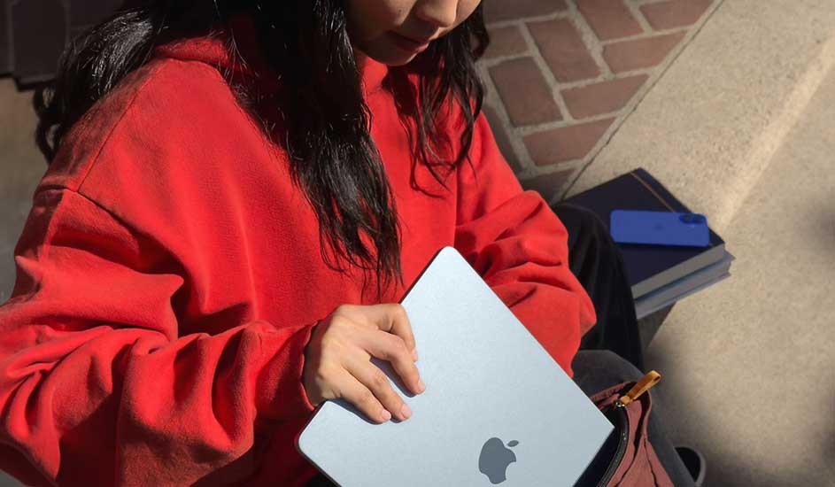 Een zilverkleurige laptop met een Apple logo wordt vastgehouden voor een bakstenen muur, naast boeken en een tas.