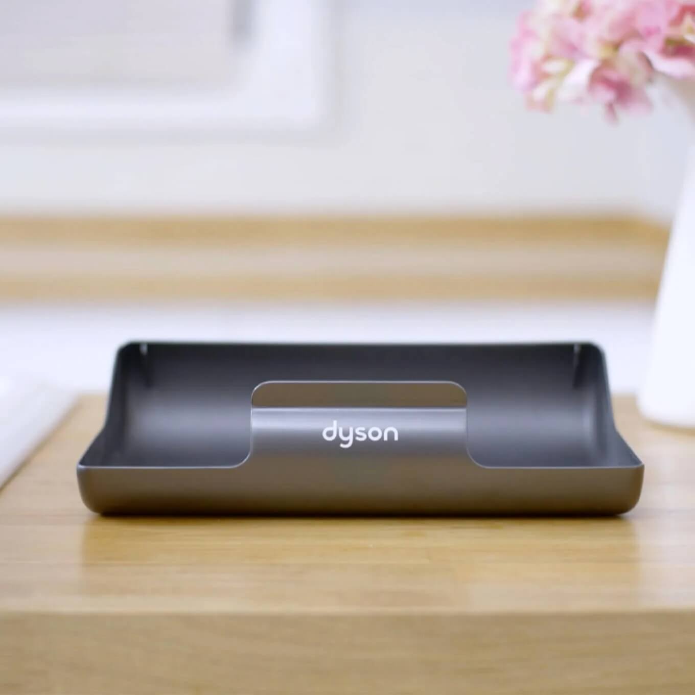 Support Dyson gris sur table en bois. Fleurs roses floues en arrière-plan.