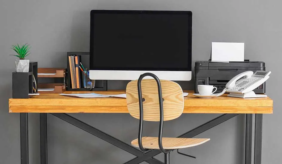 Bureau en bois avec ordinateur, imprimante, téléphone, tasse, plante, chaise sur fond gris.
