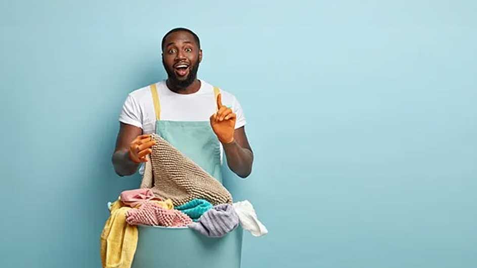 Een blauwe achtergrond toont een blauwe wasmand gevuld met kleurrijke handdoeken, met een beige schort.