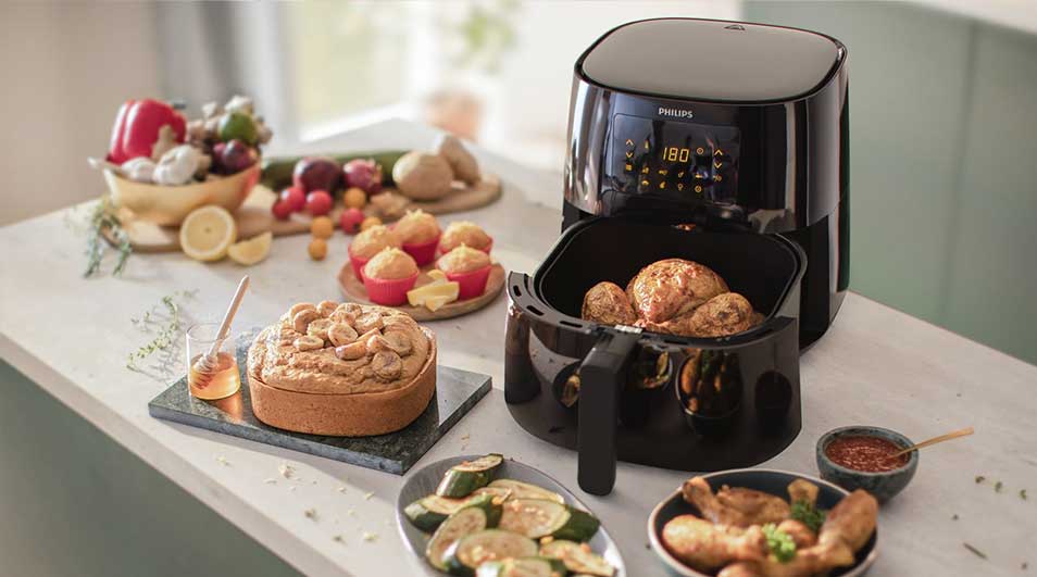 Een zwarte Philips Airfryer staat op een tafel met diverse gerechten, waaronder gebak en groenten.
