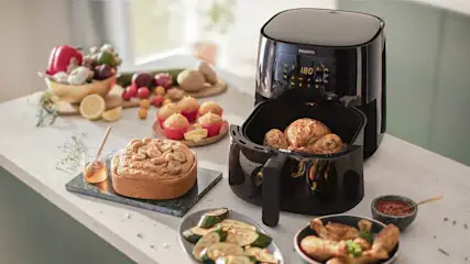 Een zwarte Philips Airfryer staat op een tafel met diverse gerechten, waaronder gebak en groenten.