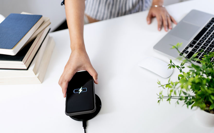Une main place un smartphone noir sur un chargeur sans fil. Un ordinateur portable, une souris, des livres et une plante sont visibles.