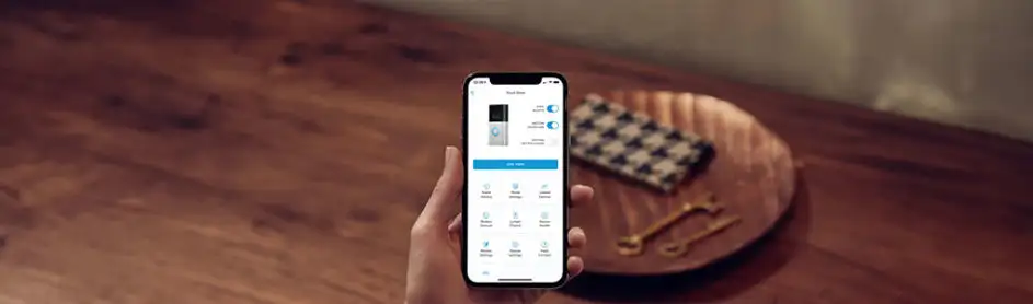 Main sur smartphone affichant une application de sonnette vidéo, près d'un plateau en bois avec un tissu et des ustensiles dorés.