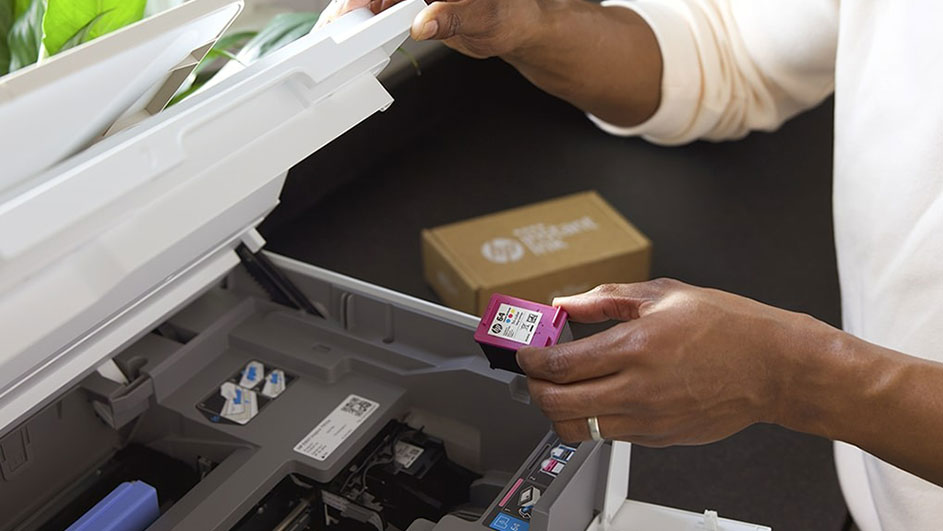 Een witte printer staat open met een roze cartridge in de hand. Een HP doos staat op de achtergrond.