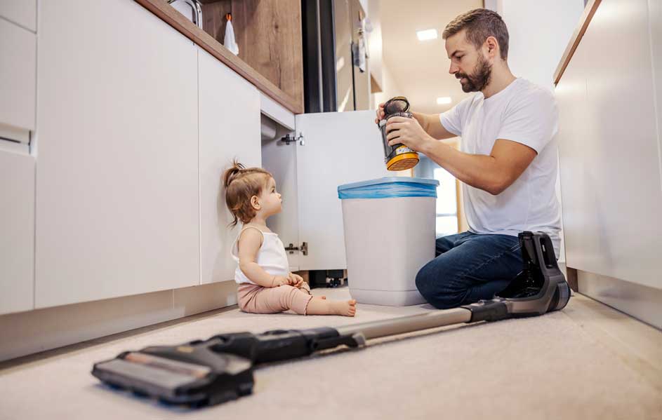 Dans une cuisine, un homme vide un récipient noir et orange dans une poubelle blanche et bleue. Un aspirateur noir repose sur le sol carrelé.