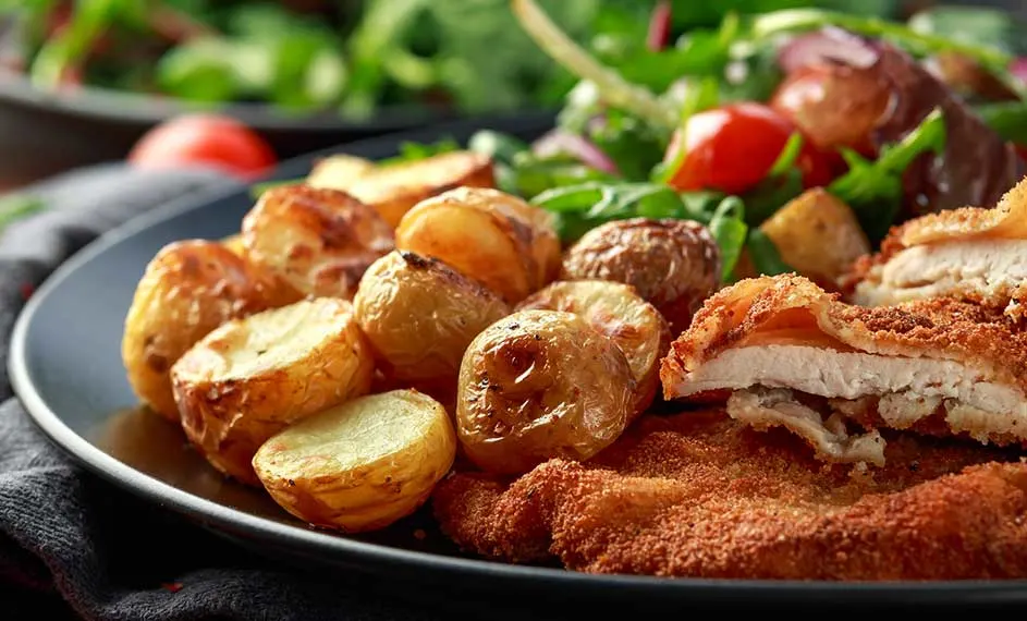 Assiette avec des pommes de terre rôties, une escalope panée et une salade verte avec des tomates cerises.