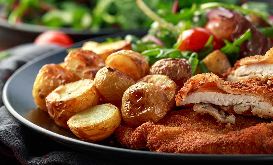 Een bord met gebakken aardappelen, schnitzel en een salade met groene bladeren en rode tomaten.