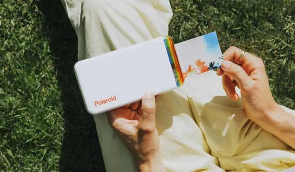 Een witte Polaroid printer rust op een schoot met geel linnen, met een foto die afgedrukt wordt tegen een groene grasachtergrond.