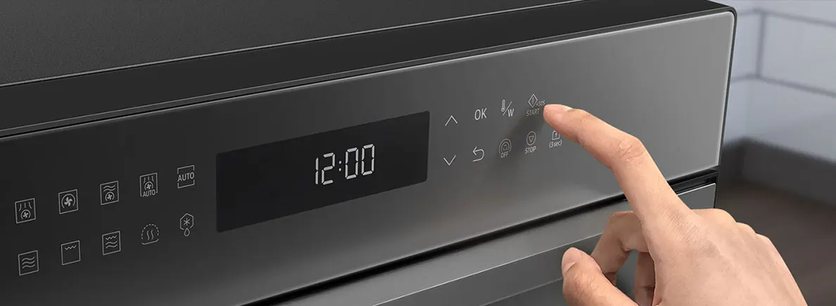 Gros plan d'un four noir avec un écran affichant 12:00. Un doigt appuie sur le bouton de démarrage.