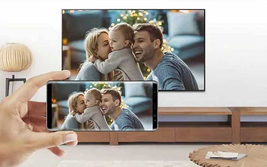 A hand holds a phone displaying a family photo, mirrored on a TV screen. The background features a wooden stand and decor.