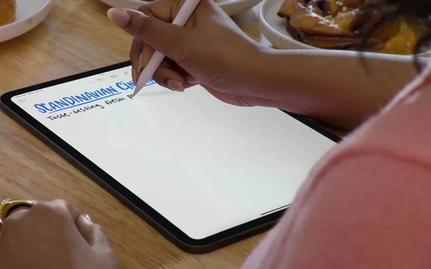 Iemand schrijft met een digitale pen op een tablet aan een houten tafel, terwijl er een bord met gebakjes naast ligt, wat een ontspannen sfeer creëert.