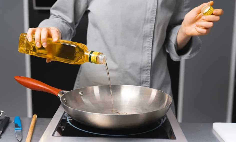 Een fles gele olie wordt in een zilverkleurige pan gegoten met een rood handvat op een zwarte kookplaat.
