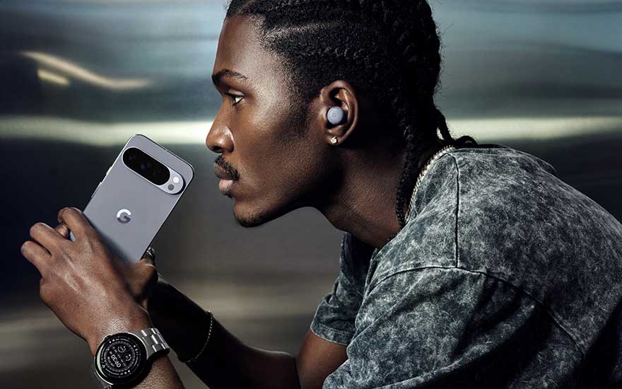 Man with dreadlocks holds grey smartphone, wearing wireless earbuds and watch in modern urban environment with soft lighting.