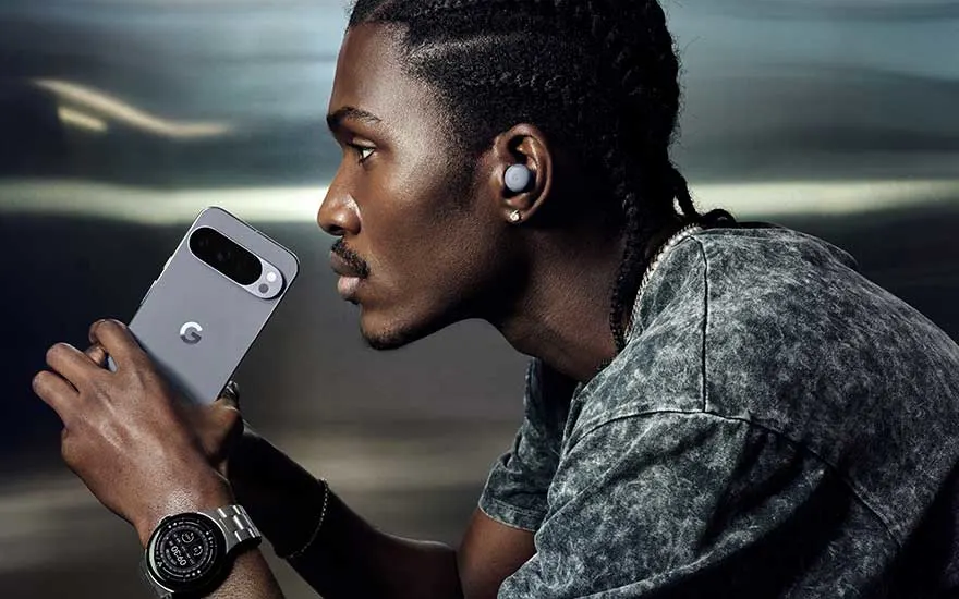Man with dreadlocks holds grey smartphone, wearing wireless earbuds and watch in modern urban environment with soft lighting.