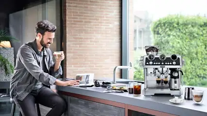 Man aan moderne keukenbar drinkt koffie en werkt op laptop naast grote zilveren espressomachine en glazen met drankjes.
