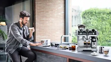 A relaxed man enjoys coffee at a sleek kitchen counter, beside a shiny espresso machine pouring two rich shots.