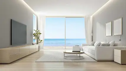 Salon lumineux avec canapé blanc, table ronde, téléviseur et vue sur la mer à travers une grande fenêtre.