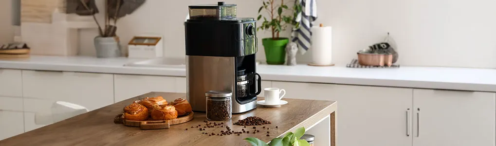Hands pour coffee beans into a grinder on a counter with a kettle and cup.