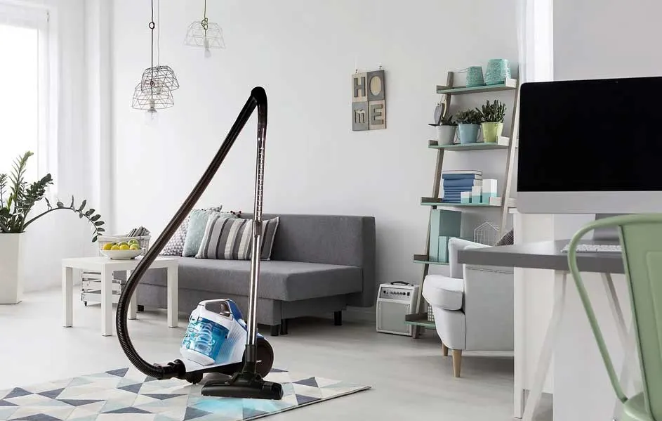 Un aspirateur bleu et blanc sur un tapis à motifs géométriques dans un salon lumineux avec canapé gris et étagère décorative.