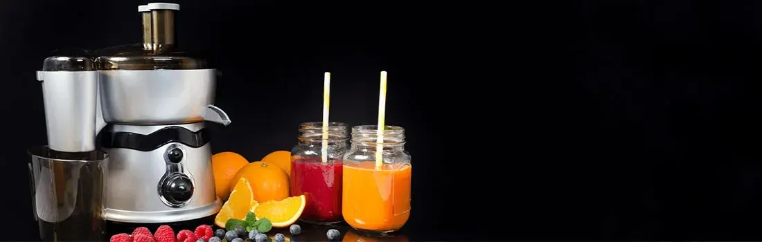 Een zilveren juicer naast sinaasappels, bessen en twee glazen gevuld met rode en oranje drankjes tegen een zwarte achtergrond.