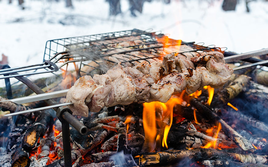 Barbecue met vlees op een spies boven een open vuur met houtblokken. De achtergrond is besneeuwd.