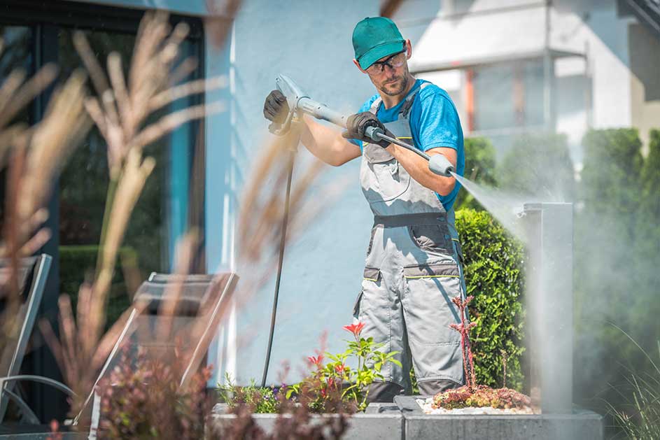 Un homme utilise un nettoyeur haute pression sur un bâtiment blanc. Des plantes sont au premier plan.