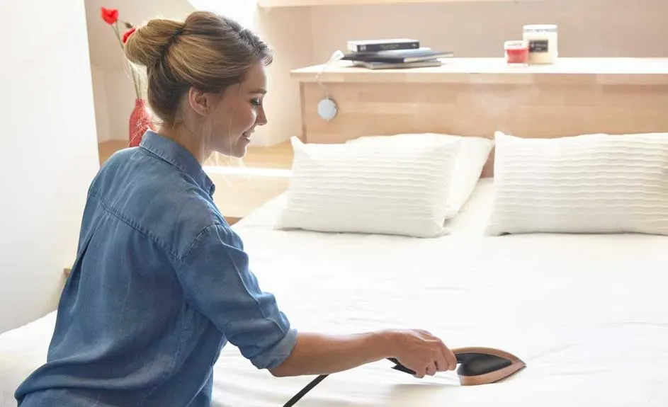 Une chambre avec un lit blanc, des oreillers, et une tête de lit en bois. Une personne utilise un appareil sur le lit.