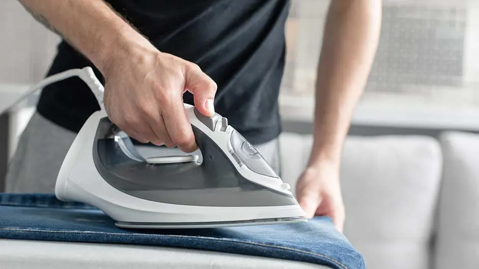 Une personne repasse un jean bleu clair avec un fer à repasser gris et blanc sur une table.
