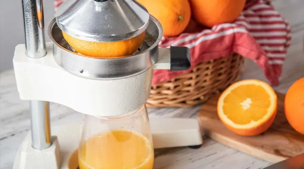 Een close-up van een sinaasappelpers met een sinaasappel erin, naast een mand met sinaasappels en sinaasappelsap in een glas.