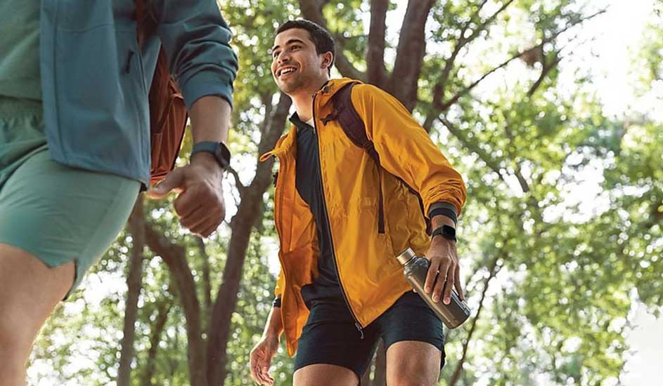 Wandelscène in bos. Man met gele jas en waterfles, vrouw met blauwgroene jas. Beide dragen een horloge.