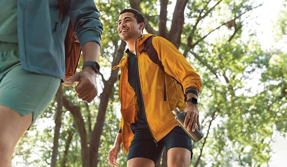 Wandelscène in bos. Man met gele jas en waterfles, vrouw met blauwgroene jas. Beide dragen een horloge.