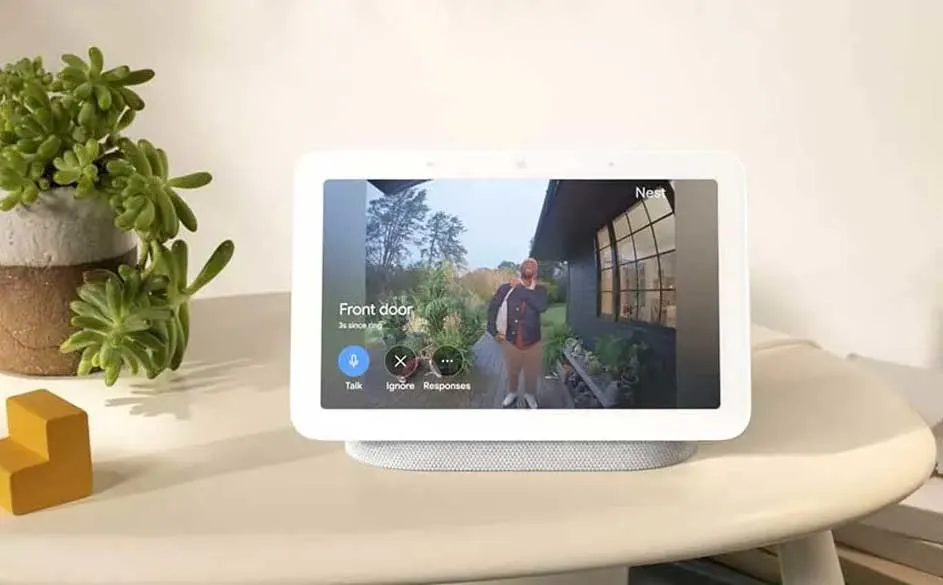 Een wit smart display toont een videofeed van een voordeur op een witte tafel. Er staat een groene plant en een geel object naast.