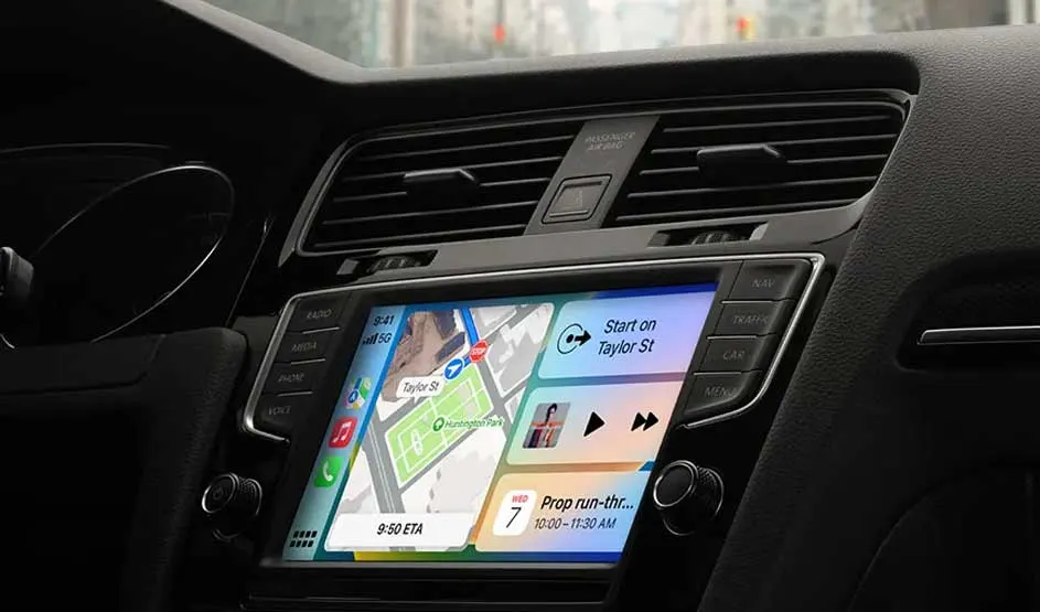 Car dashboard with Apple CarPlay on the centre screen: Maps shows a route, music and calendar tiles beside it, buttons and vents framing the display.