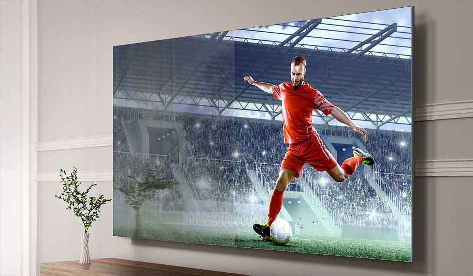 Grote tv toont voetballer in rood tenue die de bal trapt, met stadionvolk op de achtergrond en scherm verdeeld in twee helften.