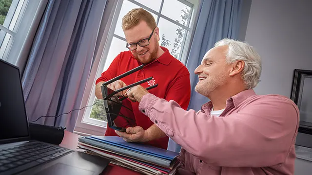 Twee mensen bij een bureau met een laptop en dossiers. Eén persoon installeert een zwarte router. Lichtinval via een raam.