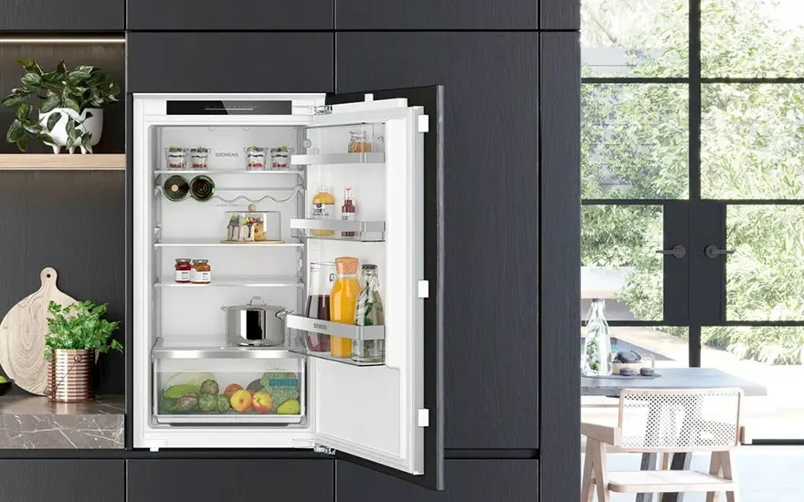 Un réfrigérateur blanc ouvert, intégré dans une armoire de cuisine sombre, montre des aliments. Une fenêtre est visible en arrière-plan.