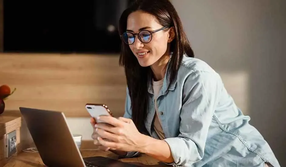 Une personne utilise un smartphone près d'un ordinateur portable sur une table en bois dans un intérieur éclairé.