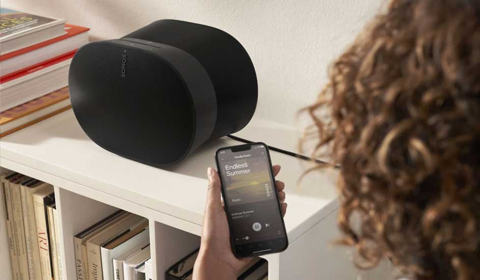 Deux enceintes noires Sonos sur étagère blanche avec livres. Un smartphone affiche "Endless Summer".