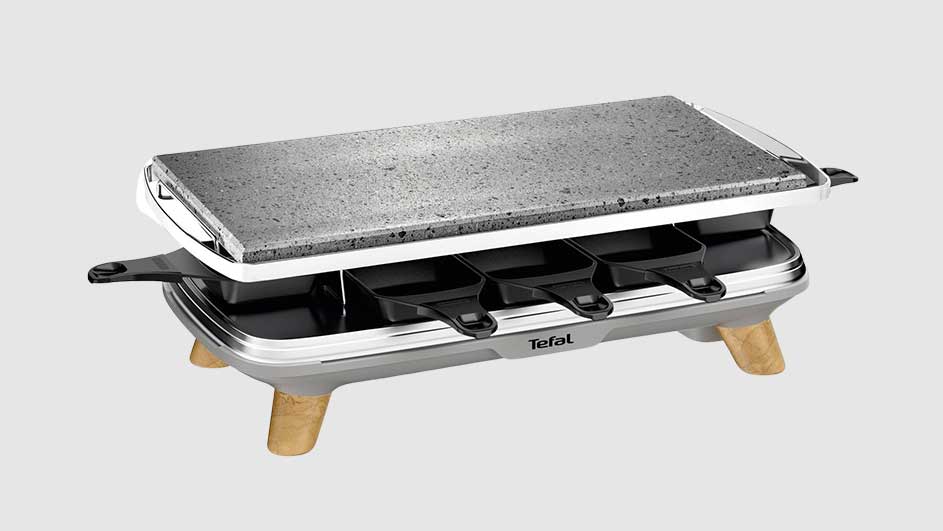Een raclette-apparaat met een grijze steenplaat bovenop, zwarte pannetjes eronder en houten poten.